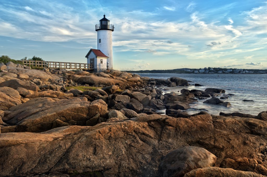 A lighthouse in the distance in Gloucester, MA with rocks in the foreground